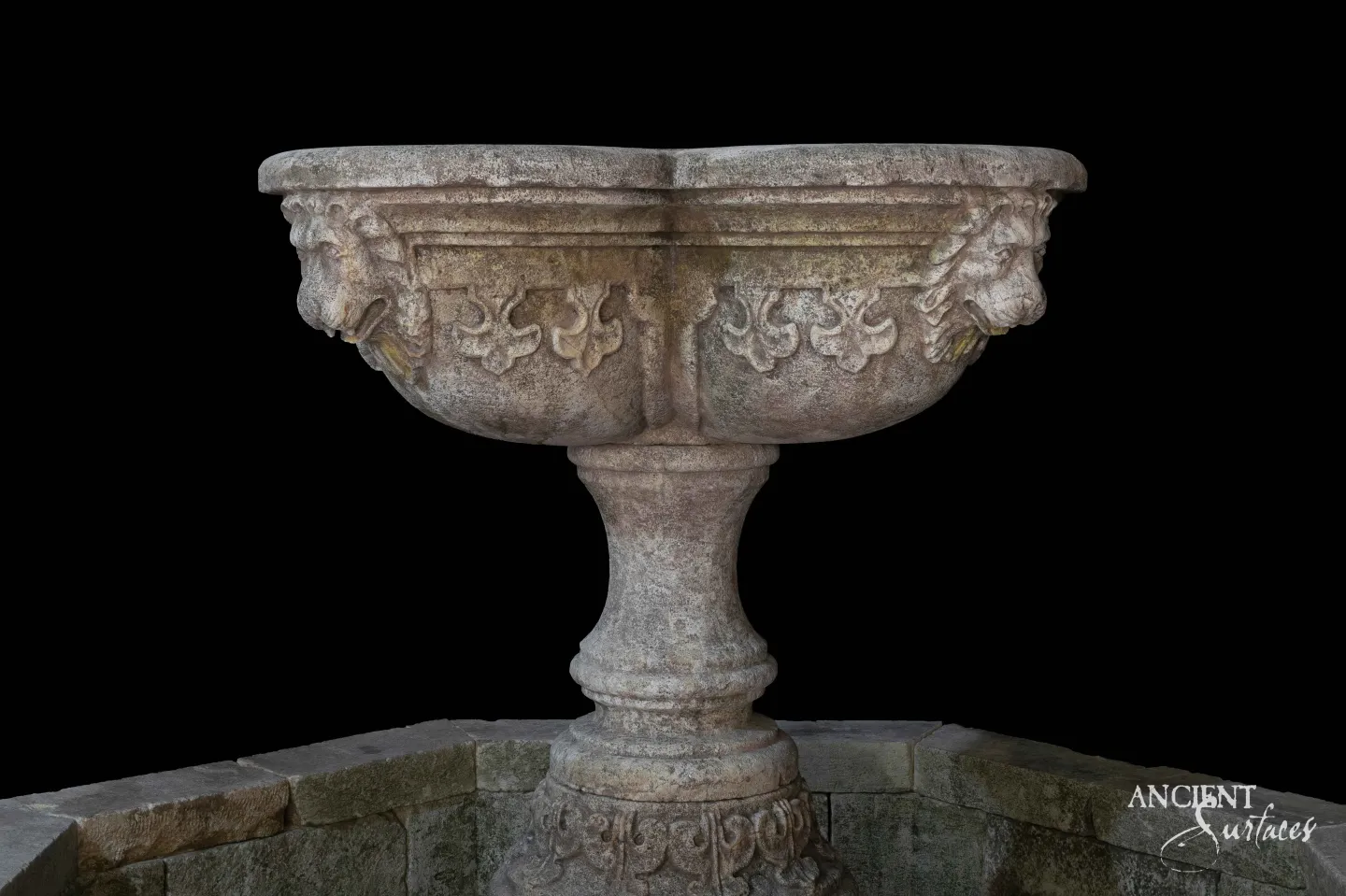 Close view of a reclaimed antique limestone fountain bowl from Ancient Surfaces carved with a band of palmette relief and lion masks with metal spouts at each corner, raised on a waisted baluster foot above rough stone coping