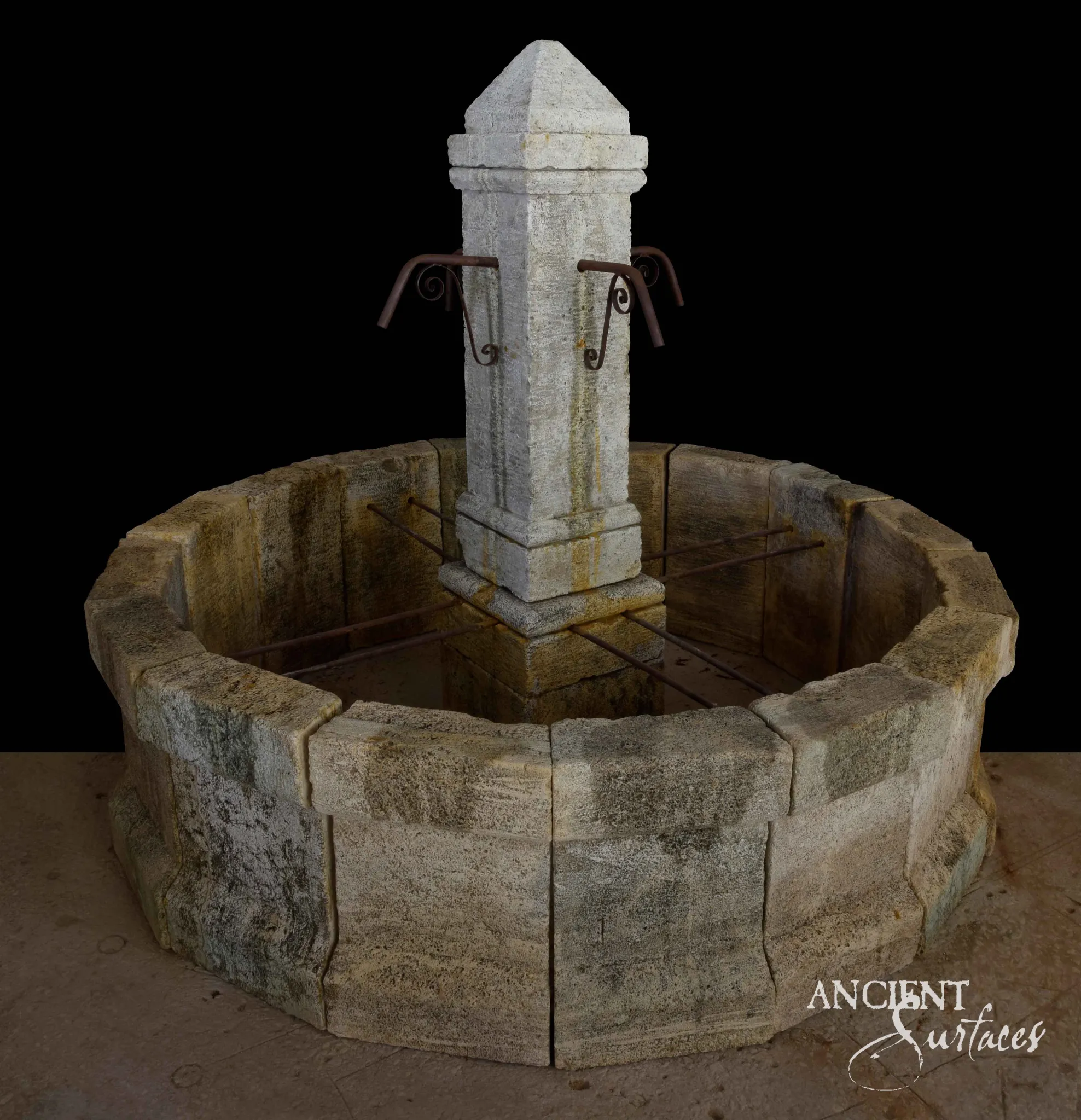 Reclaimed antique limestone pool fountain from Ancient Surfaces with a round basin of thick rough faced blocks heavily grown with green and ochre lichen, and a pale tapered central pillar with a pyramidal cap and iron scroll spouts