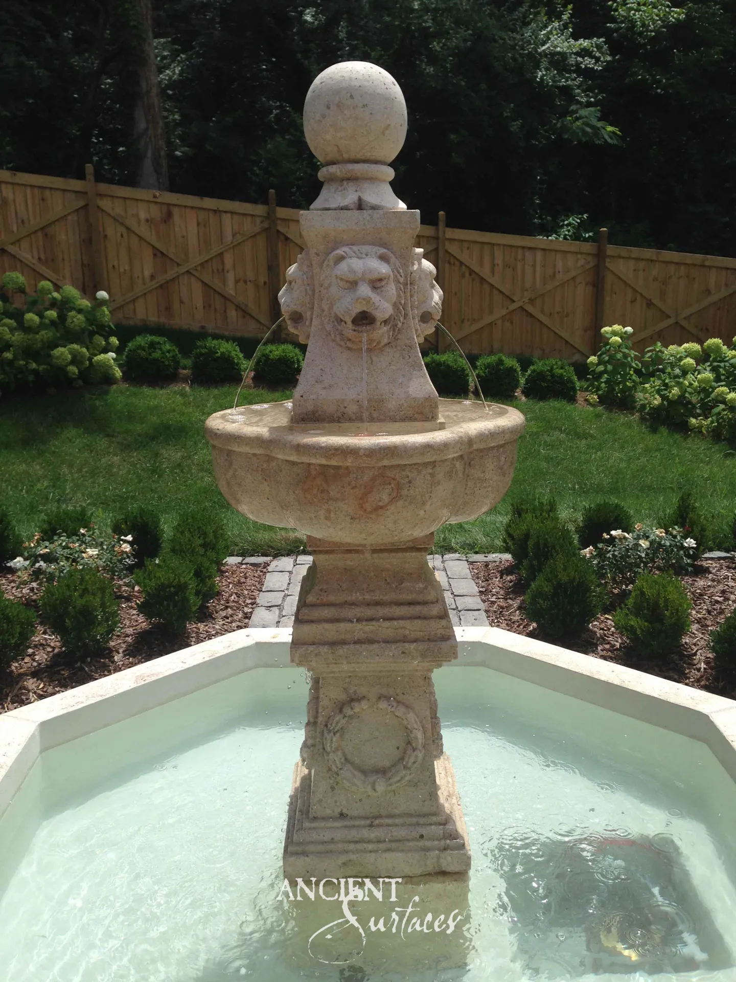 Reclaimed antique limestone pool fountain from Ancient Surfaces running in a planted garden pool, a carved lion mask spouting three arcs of water into a scalloped bowl above a pedestal carved with a laurel wreath