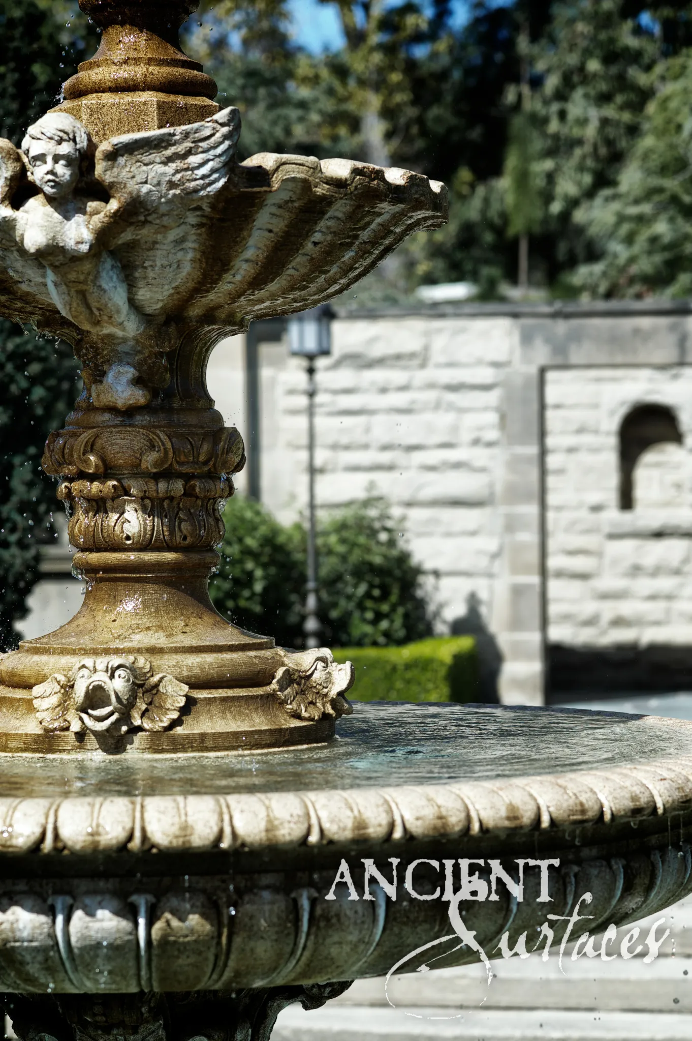 Close view of a running reclaimed antique limestone fountain from Ancient Surfaces, a winged cherub carved beneath a scalloped shell bowl above an acanthus collar and grotesque masks at the foot of the stem