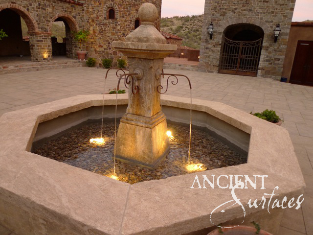 Reclaimed antique limestone pillar pool fountain from Ancient Surfaces lit at dusk in a stone courtyard, four falls of water from wrought iron scroll spouts dropping into a shallow octagonal pool laid with river pebbles